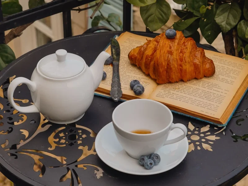 Mesa pequeña de exterior con taza, tetera y un cruasán junto a plantas