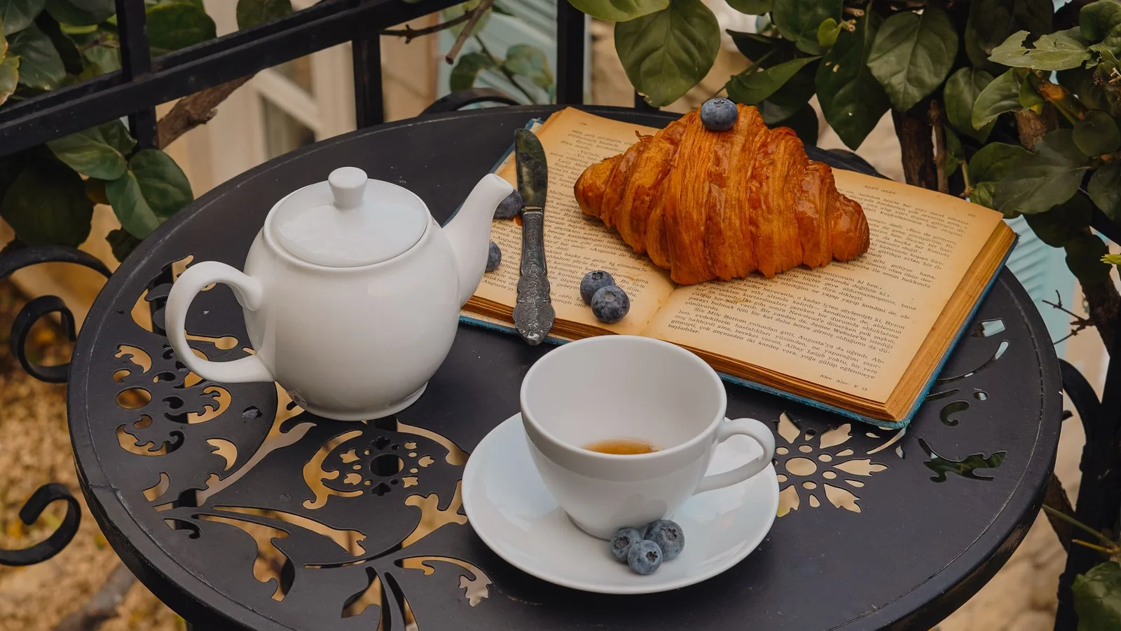 Mesa pequeña de exterior con taza, tetera y un cruasán junto a plantas