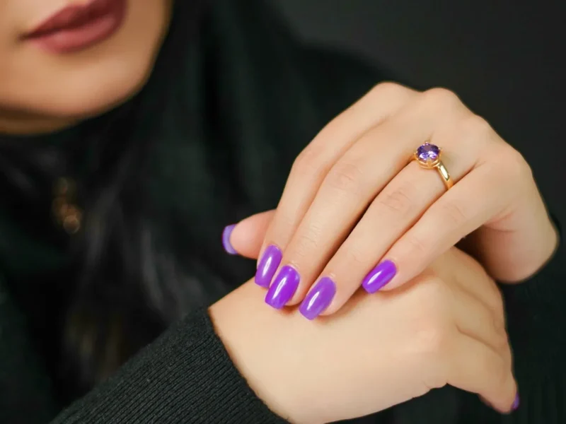 Manos con uñas de color violeta y un anillo delante de una prenda negra.
