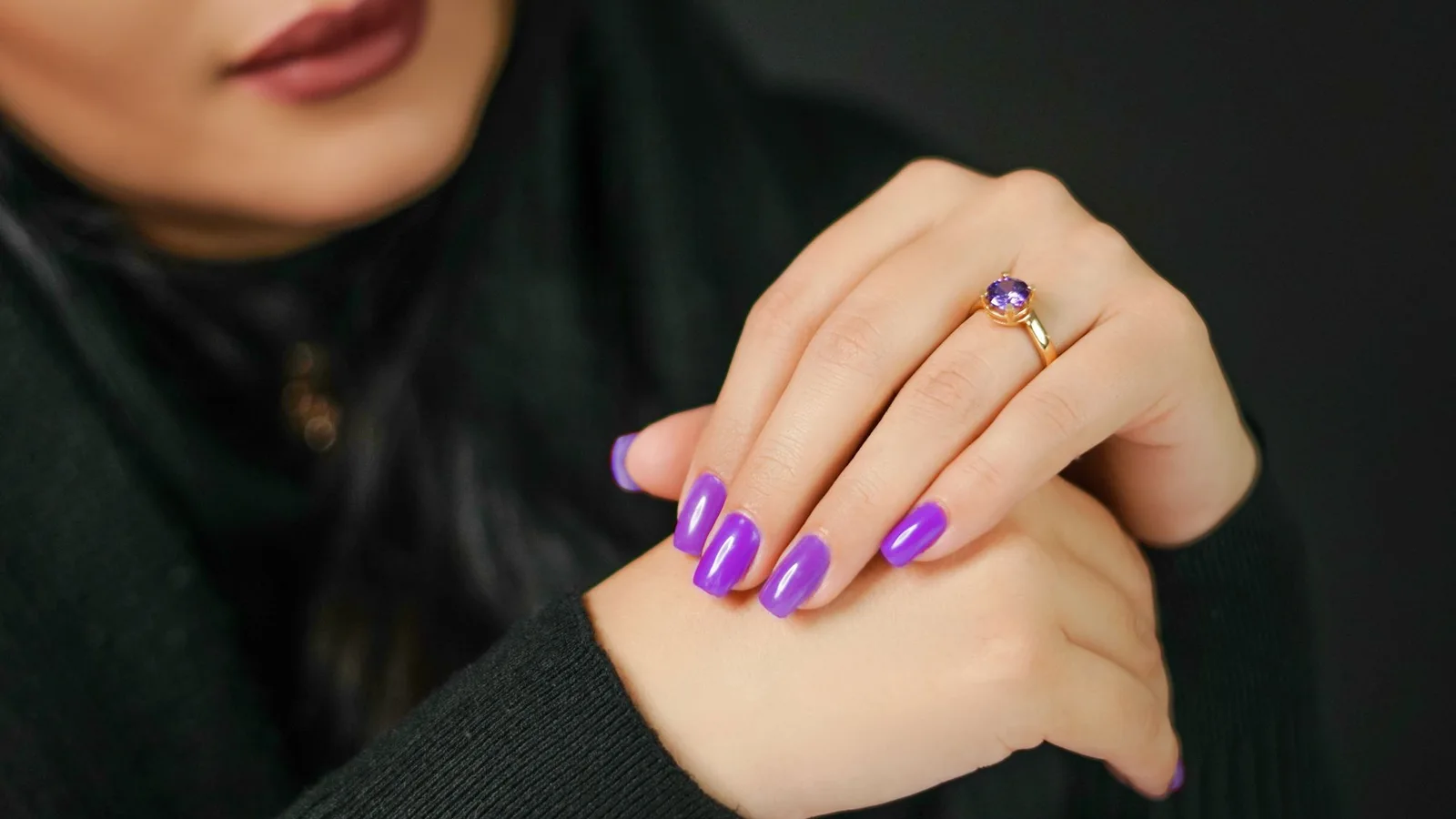 Manos con uñas de color violeta y un anillo delante de una prenda negra.