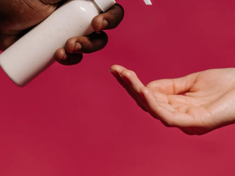 Manos sujetando y accionando un envase cosmético blanco con dosificador sobre fondo rosa.