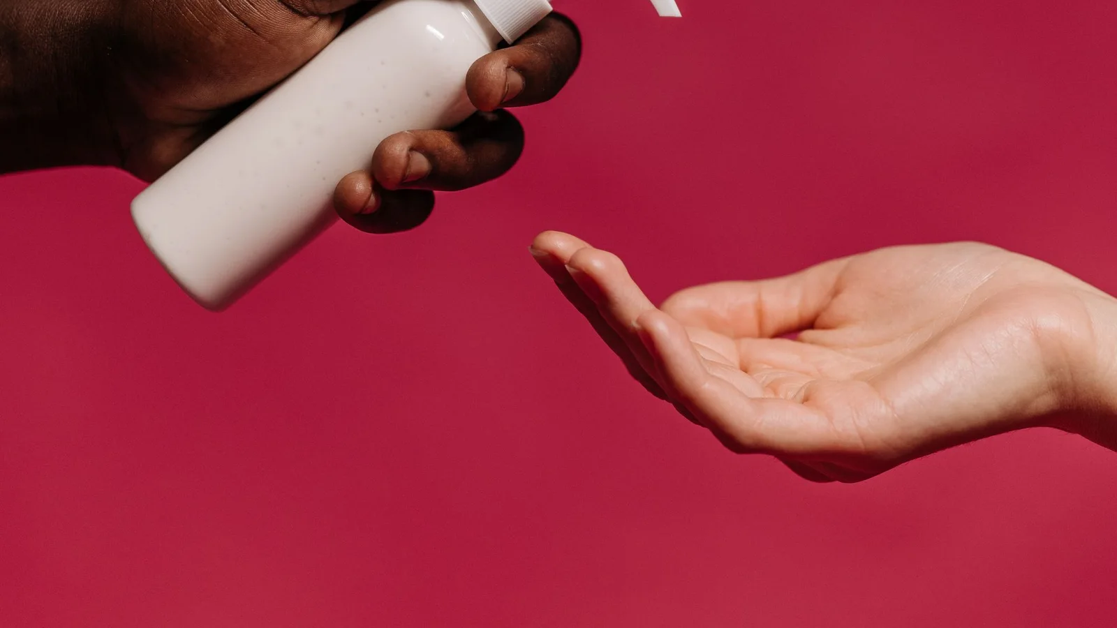 Manos sujetando y accionando un envase cosmético blanco con dosificador sobre fondo rosa.