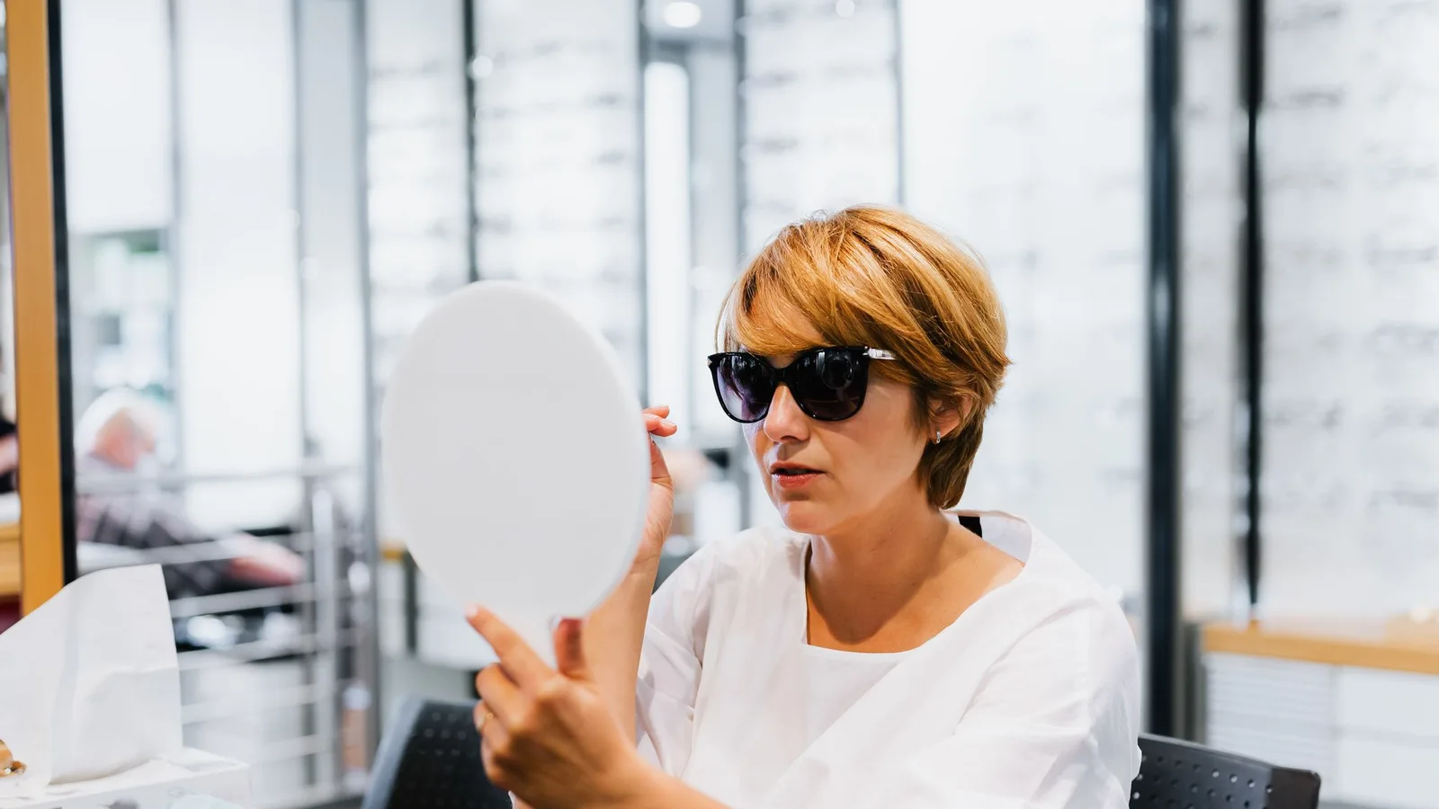 Mujer probándose unas gafas de sol y observándose en un espejo de mano dentro de una óptica.