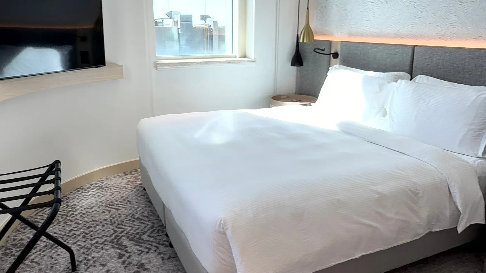 Habitación con cama blanca, ventana y soporte para equipaje