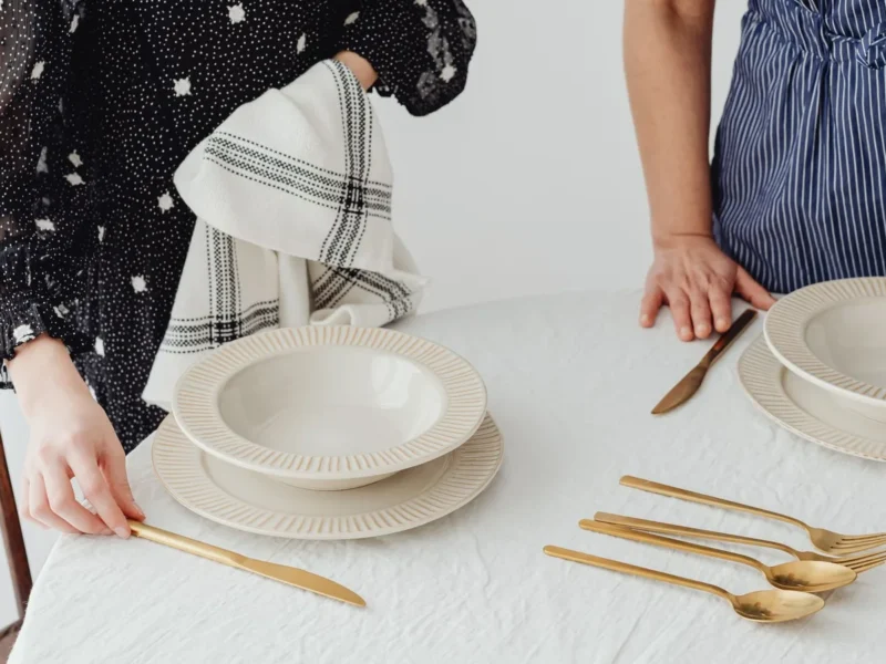 Dos personas colocando platos claros y cubiertos sobre una mesa con mantel