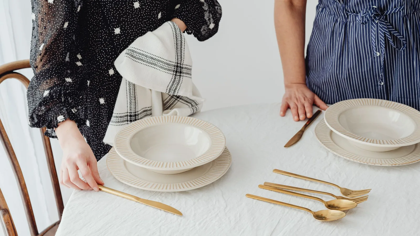Dos personas colocando platos claros y cubiertos sobre una mesa con mantel