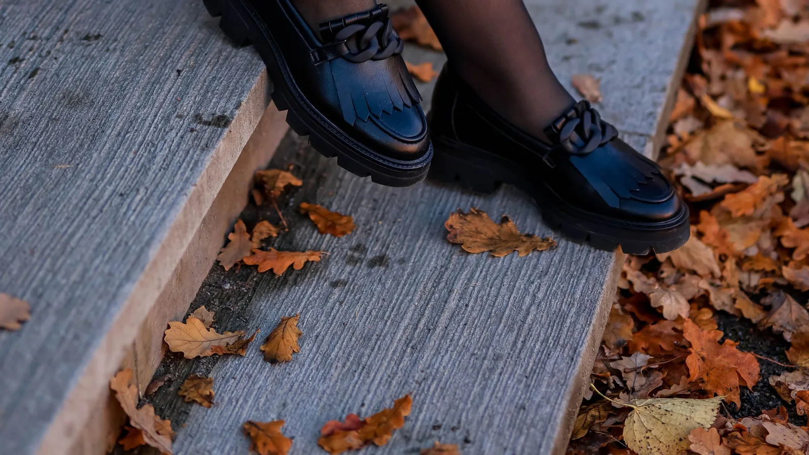 Mocasines negros con detalle metálico junto a unas hojas caídas