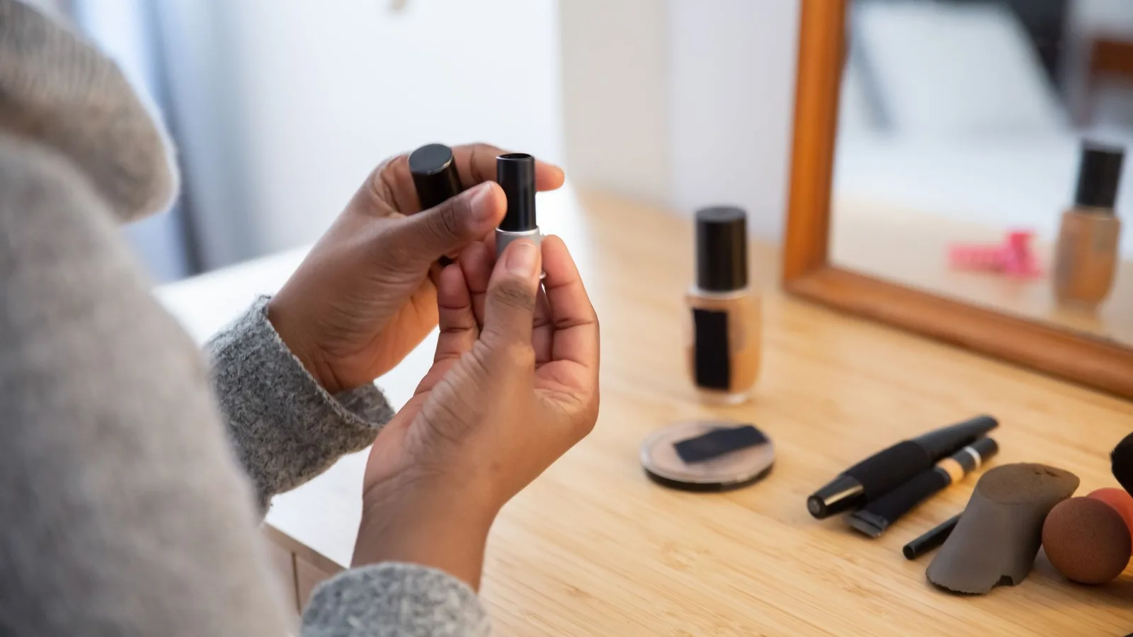 Manos examinando una barra de labios junto a cosméticos colocados delante de un espejo.